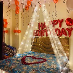A classic romantic birthday setup with a fairy light canopy over the bed. The room is decorated with red and white balloons, and a heart of rose petals on the bed adds a special touch.