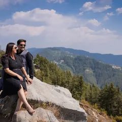 The couple sits together on a rock, looking out at the expansive green and blue mountain ranges. A perfect moment of shared peace and admiration for the view.