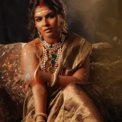 A wider shot showing the model seated on rocks, grounding the divine styling in an earthy, raw environment. The flowing tissue saree contrasts with the rugged setting.