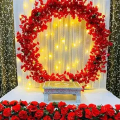This modern Ganpati decoration features a bold circular ring completely covered in vibrant red artificial flowers. Set against a curtain of fairy lights, this design provides a contemporary and striking focal point for your celebration.