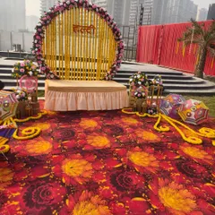 A wide view of an outdoor Haldi setup, showing the main stage with the floral backdrop, Rajasthani umbrellas, and a colorful printed carpet.