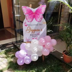 A beautiful butterfly-shaped welcome sign for a first birthday party. The sign is decorated with pink and white balloons and set up at the entrance to greet guests.