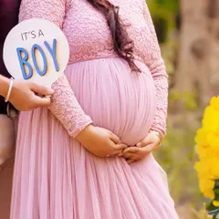 A close-up shot focusing on an "It's a BOY" sign held next to the baby bump. The soft pink of the gown and the outdoor setting create a lovely gender reveal photo.