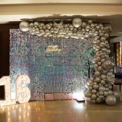 A dazzling setup for a 16th birthday party. The iridescent sequin wall creates a stunning, shimmering backdrop, complemented by a silver balloon garland and large marquee numbers.