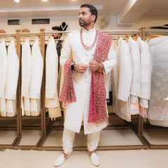 A full-length view of a groom trying on a classic ivory sherwani, paired with a rich red embroidered stole. Notice how the outfit looks against our in-store collection.