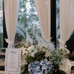 An elegant table centerpiece from a blue Dior-inspired shower. A giraffe figure stands amidst a lush arrangement of blue and white hydrangeas and baby's breath, next to a custom menu card.