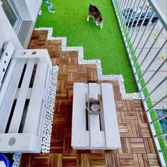 This balcony is now a chic and pet-friendly outdoor lounge. The design features a mix of wooden deck tiles and artificial grass, bordered with white pebbles, and furnished with custom pallet seating.