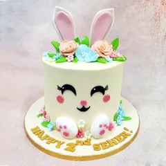 We craft pure pastel joy with this adorable bunny face cake. Perfect for a second birthday, this design features a sweet bunny face with large ears peeking out from a crown of delicate, handcrafted sugar flowers in soft peach and blue.