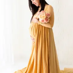 A full-length portrait of a mother-to-be in a long yellow gown, holding a bouquet of flowers. The soft, natural light from the window enhances the gentle mood of this boho session.
