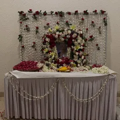 Another memorial setup with a beautiful floral wreath around the photograph. The table is decorated with a white cloth, scattered petals, and a garland of jasmine flowers.