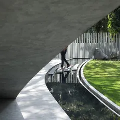 A unique view from beneath the villa's cantilevered form, showing a person crossing a water feature on stepping stones. This highlights the intimate connection between the architecture and the curated landscape.