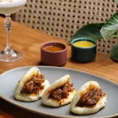 Showcasing these delicious pork bao buns with their accompanying dips. The composition is balanced with a refreshing cocktail to present a complete and satisfying meal option.