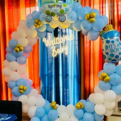 A classic "Welcome Baby" boy decoration. This setup features a circular balloon arch in blue and white, a neon sign, and foil balloons shaped like a baby carriage and a milk bottle.