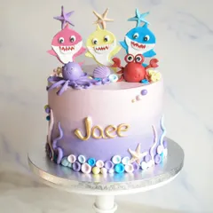 A vibrant Baby Shark cake for baby Jaee's second birthday. This design features a purple ombre effect and is decorated with handmade fondant sea creatures like an octopus and crab, along with the Baby Shark family.