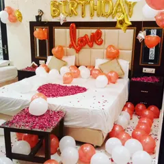 A bright and cheerful birthday room setup with rose gold, white, and gold balloons. The decor features 'Happy Birthday' and 'Love' foil balloons, and a rose petal heart on the bed.
