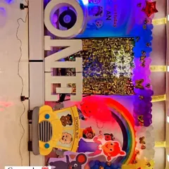 This Cocomelon themed first birthday setup features a 'ONE' marquee sign and a backdrop with a rainbow and school bus cutout. The balloon arrangement adds a playful touch to the celebration.
