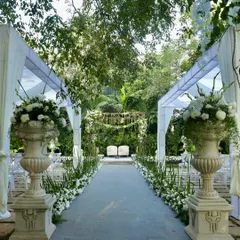 A pristine white and green aisle, flanked by large classical urns filled with white flowers, leading to a garden mandap.