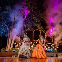 A high-energy sangeet performance by the bride and her best friend. The stage is set with dramatic smoke effects and purple uplighting against a backdrop of trees and illuminated arches, creating a magical forest vibe.