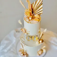 A sophisticated two-tier cake for a 60th birthday celebration in an ivory and gold theme. This design features a clear separator tier, macarons, and dried palm leaves for a modern, celebratory feel.