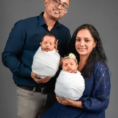 A classic posed portrait of a family with their newborn twins. The parents smile at the camera while securely holding their sleeping babies, creating a perfect first family photograph.