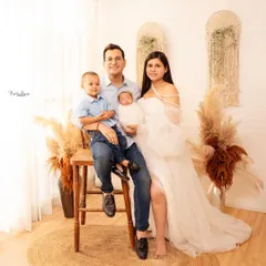 This portrait captures a relaxed family moment. The father holds their older son while the mother holds the newborn, all coordinated in a light and airy color palette against a boho-chic backdrop.
