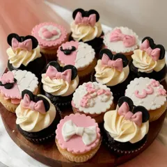 A collection of Minnie Mouse themed cupcakes for a third birthday party. The assortment includes vanilla and chocolate cupcakes with buttercream swirls topped with fondant Minnie ears and pink bows, as well as flat fondant toppers with the number '3'.