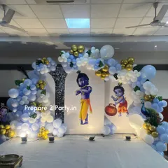 A grand and traditional Krishna-themed decoration for a naming ceremony or birthday. The stage features cutouts of Little Krishna against backdrop panels, framed by blue, white, and gold balloon arches and a beautiful peacock prop.