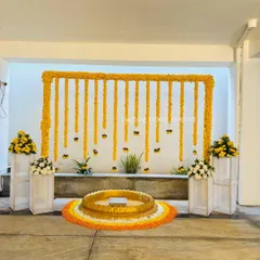 A clean and modern Haldi setup with a marigold arch against a white wall. The large brass urli is placed on a rangoli made of flower petals, combining traditional and contemporary elements.