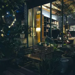An evening view of our studio's reading nook and library, seen from the garden. The warm light from within spills out into the landscape, highlighting the seamless transition between the interior and the plant-filled exterior.