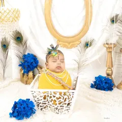 A serene portrait of a baby dressed as little Krishna, sleeping peacefully in a white basket. The setup includes peacock feathers and royal blue flowers for a divine touch.
