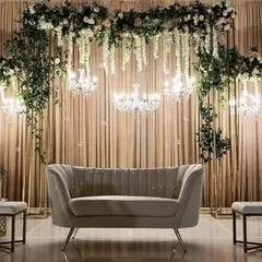 A chic and modern engagement stage setup. The design features a backdrop of cascading florals, elegant chandeliers, and a stylish grey sofa, perfect for a sophisticated celebration.