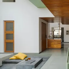 A sunken seating lounge with low, concrete-base furniture and bright yellow cushions creates an informal and cozy gathering spot. The design uses varying floor levels to define zones without walls, maintaining an open and airy feel.