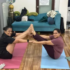 Two students working together in a supported boat pose, demonstrating the community and connection we foster in our classes.