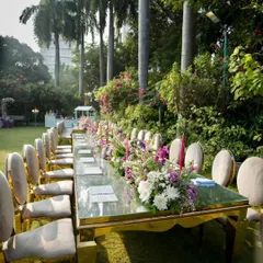 A full length view of the grand dining table setup. The gold-framed chairs and mirror-top table reflect the beautiful garden surroundings and floral arrangements.
