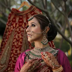 A profile shot capturing the elegance of a bride on her wedding day. This look highlights a sharp contour, a classic bridal bun, and intricate jewelry. The makeup is designed to be long lasting and picture perfect from every angle, with her bridal lehenga hanging in the background, ready for the ceremony.