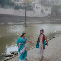 Walking along the ghats, hand in hand. This wide shot captures the couple in their beautiful traditional attire against the backdrop of the river and old city buildings.
