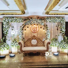 An elegant stage setup combining a circular floral arch with delicate peacock feather accents. The design uses a mix of blue and white flowers, complemented by a custom monogram and a classic gold and cream sofa.