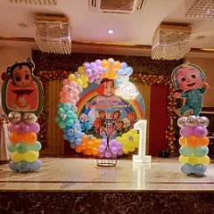 A colorful Cocomelon theme for a first birthday party, set up on a stage. This includes a circular backdrop with the baby's photo, character cutouts, a rainbow balloon arch, and a light-up number "1".