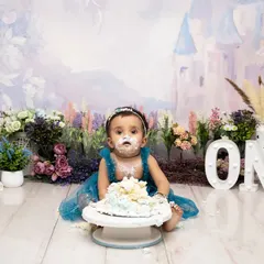 The aftermath of a successful cake smash. This little one is covered in blue frosting from head to toe, sitting proudly in front of her demolished cake in a magical floral setting.