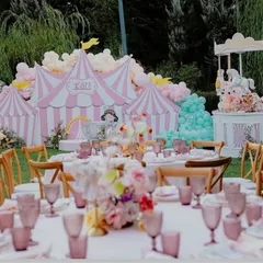 A dreamy carousel-themed party in a beautiful garden setting. The view from the guest tables shows the grand circus tent backdrop and carousel cake station, creating a picturesque scene for a special birthday.