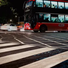 A classic red double-decker BEST bus crosses a zebra crossing in Mumbai at night. The long exposure shot captures the motion of a passing car as a white streak, contrasting with the stationary bus.