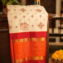 This stunning white Kanjivaram features intricate Kasuthi embroidery from Karnataka, with elephant and geometric motifs, on a bold red and orange gap border.