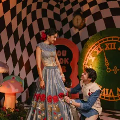 A romantic moment in Wonderland. The groom adjusts the bride's rose-adorned lehenga against a surreal checkered backdrop, a perfectly staged shot within the fantastical theme I designed for their Mehndi night.