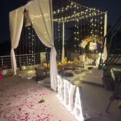 A romantic "Bride to Be" terrace decoration. The setup includes a canopy with white drapes and fairy lights, scattered rose petals, and large marquee letters for the name.