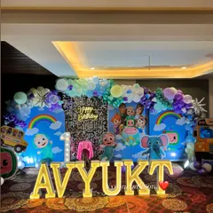 For this grand first birthday, I created a full Cocomelon wonderland. The setup includes a massive backdrop with characters, a silver sequin panel, a balloon arch, large light-up marquee letters spelling the name 'AVYUKT', and multiple character cutouts.