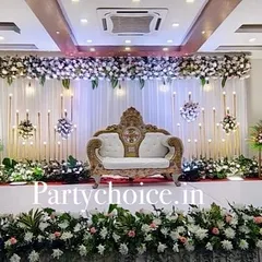 Here is an elegant indoor reception stage we designed. It features a classic white floral theme with a beautifully carved sofa for the couple, a clean white backdrop with hanging lights, and floral arrangements on pedestals.