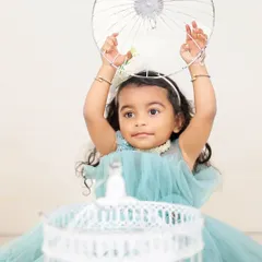A young girl in a light blue dress plays with a birdcage prop during an outdoor session. The natural background and soft lighting create a dreamy and ethereal portrait.