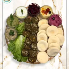 A beautifully arranged Mezze Platter on a round wooden board. This version features crispy falafel, mini pitas, and a colorful array of five different dips, including hummus and tzatziki.