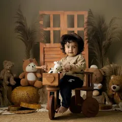 A toddler in a cute hat sits on a wooden tricycle, surrounded by teddy bears in a warm, rustic studio setting.