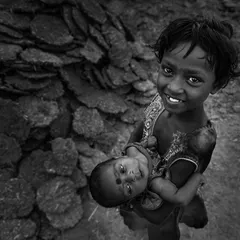 A young girl smiles warmly as she holds her infant sibling, a heartwarming black and white portrait of love and responsibility in a rural setting.
